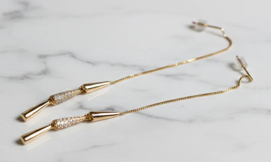 Orecchini pendenti dorati con dettagli brillanti su sfondo in marmo bianco, gioielli eleganti.